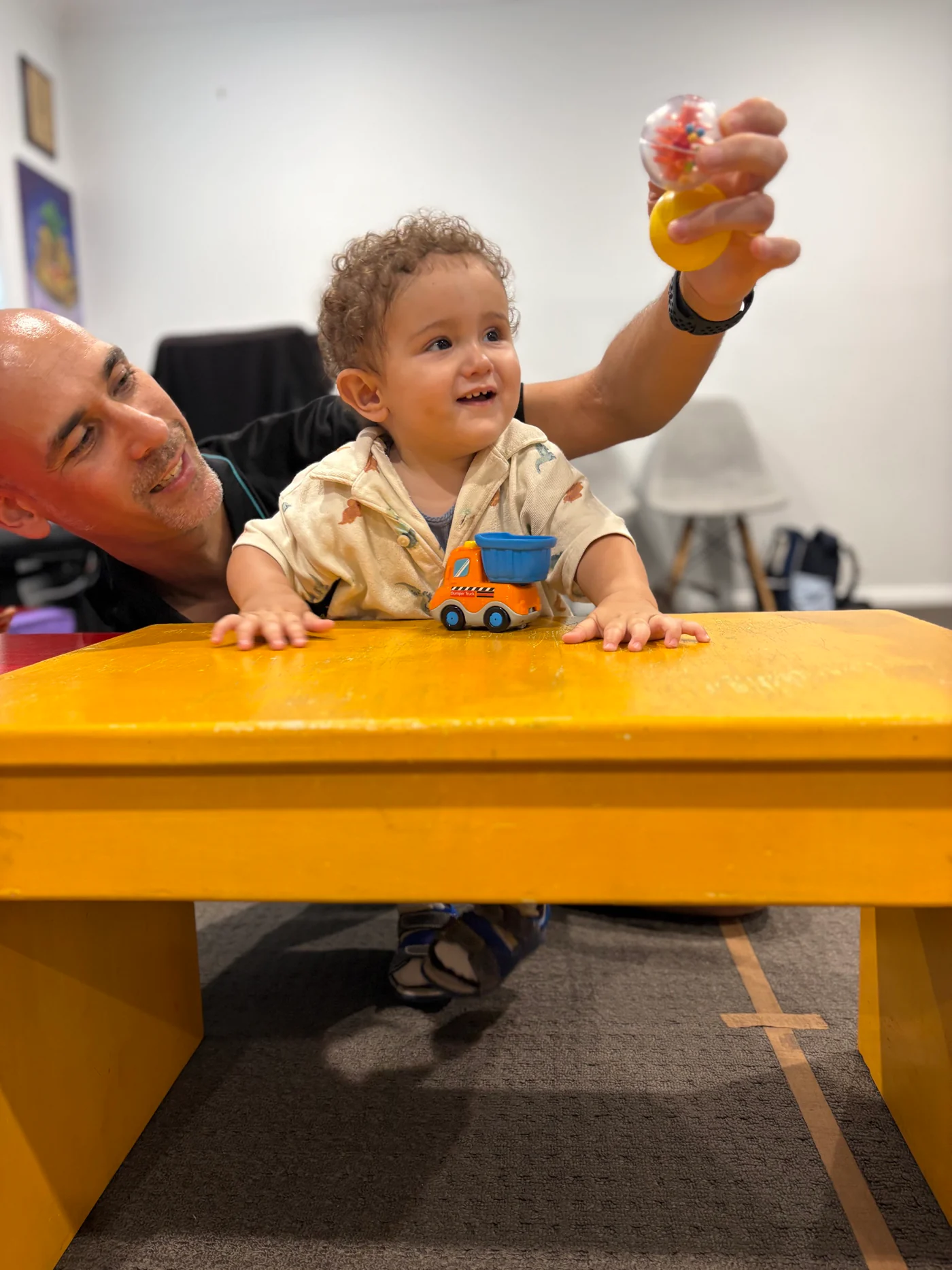A physio helping a toddler stand at a table and reach for a ball